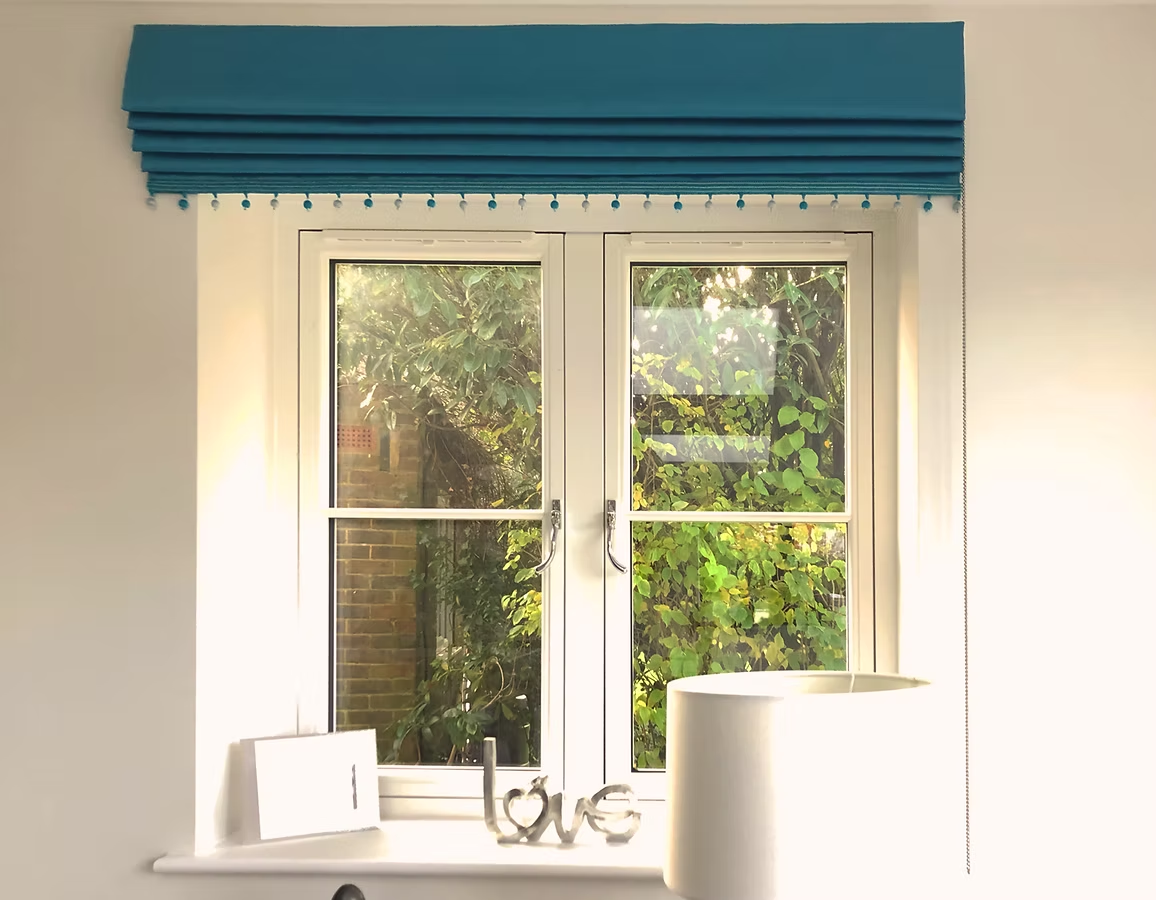 Teal Roman blind finished with beaded trim above a cottage-style window