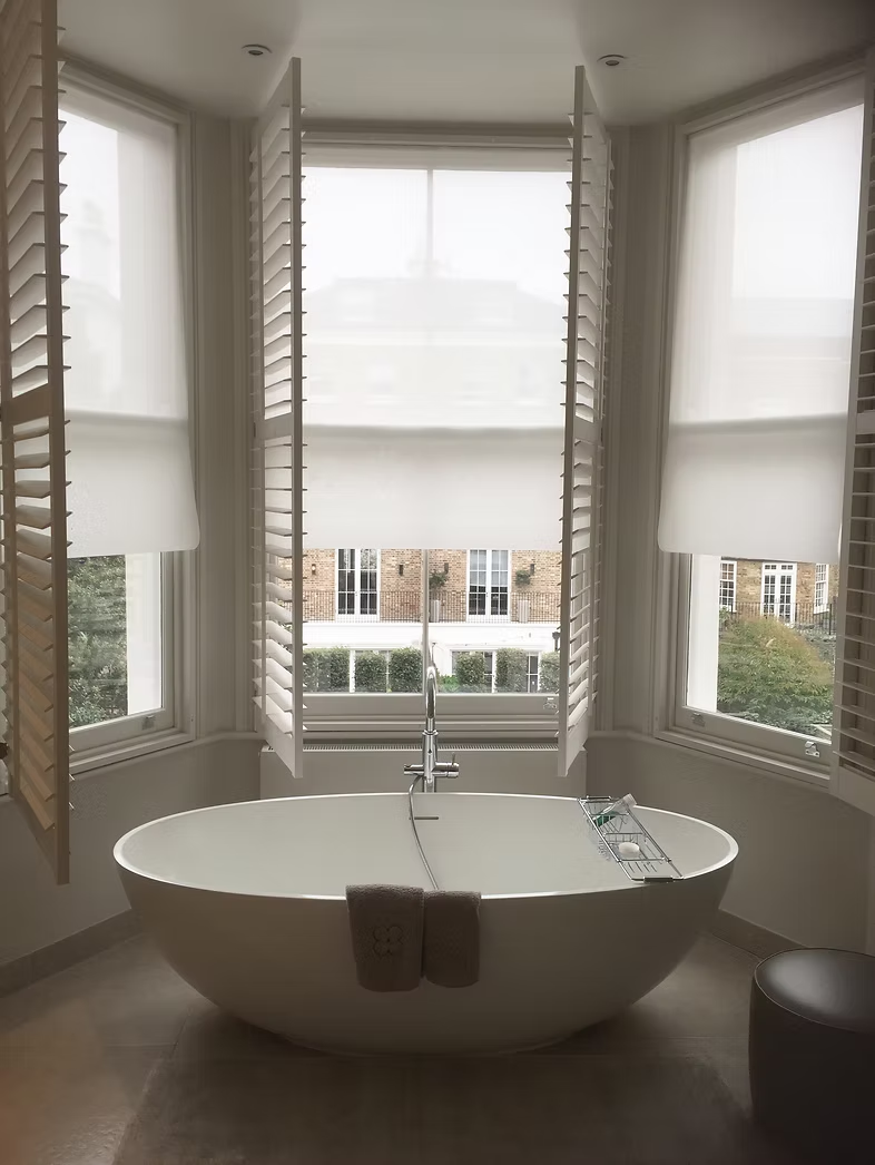Plantation shutters and white roller blinds surrounding a freestanding bath