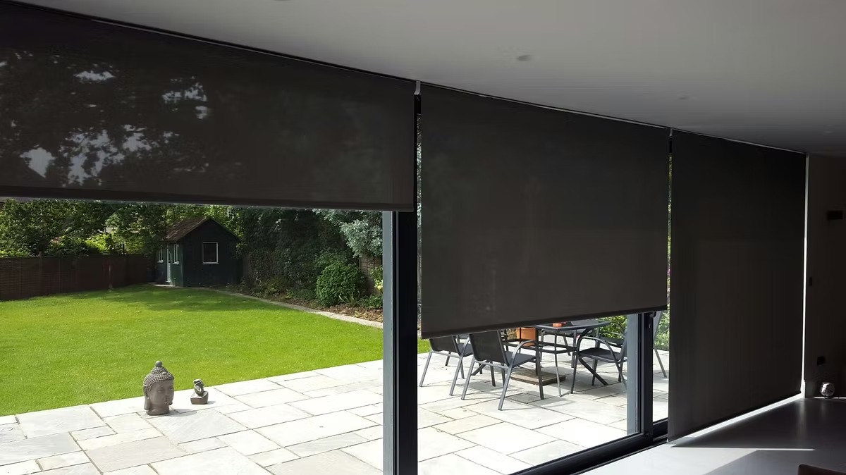 Dark motorised roller blinds covering wide glazed garden doors onto a patio and lawn
