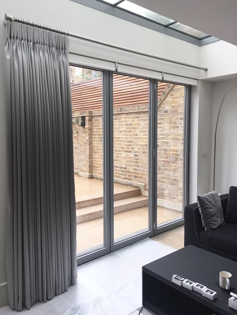 Tall grey bespoke curtains hung beside bi-fold doors under a glazed roof